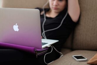 Woman on an apple laptop with earbuds plugged into the iphone beside her on the couch.