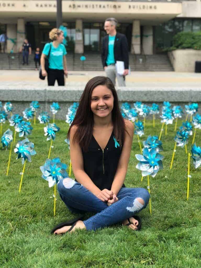 Former gymnast Kaylee Lorincz sits outside the Hannah Administration Building on Michigan State University campus in 2018 surrounded by teal pinwheels placed to represent support for survivors of ex-MSU and Olympic doctor Larry Nassar.