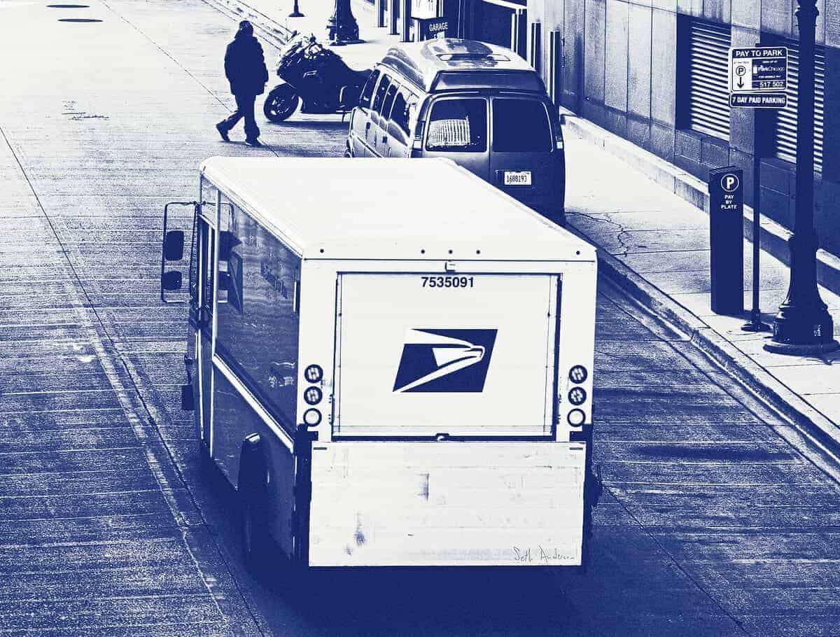 A black and white photo of a usps truck driving down a city street. Viewed from above and behind.