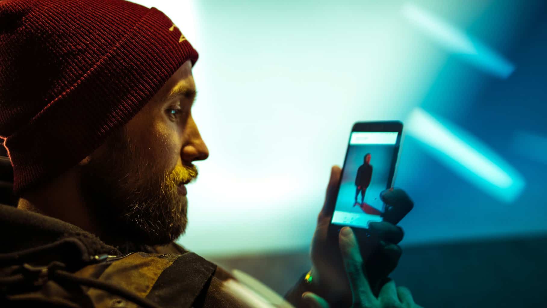 Become less visible online. A man with a beard, wearing a beanie, watching a video on his cell phone.