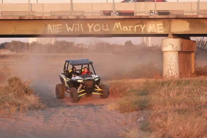 An ATV driving through the dirt by the Arkansas river under a bridge with “Will you marry me” scrawled in the dirt