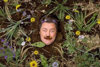 Comedian mimics growing out of the dirt with only his face showing surrounded by wildflowers