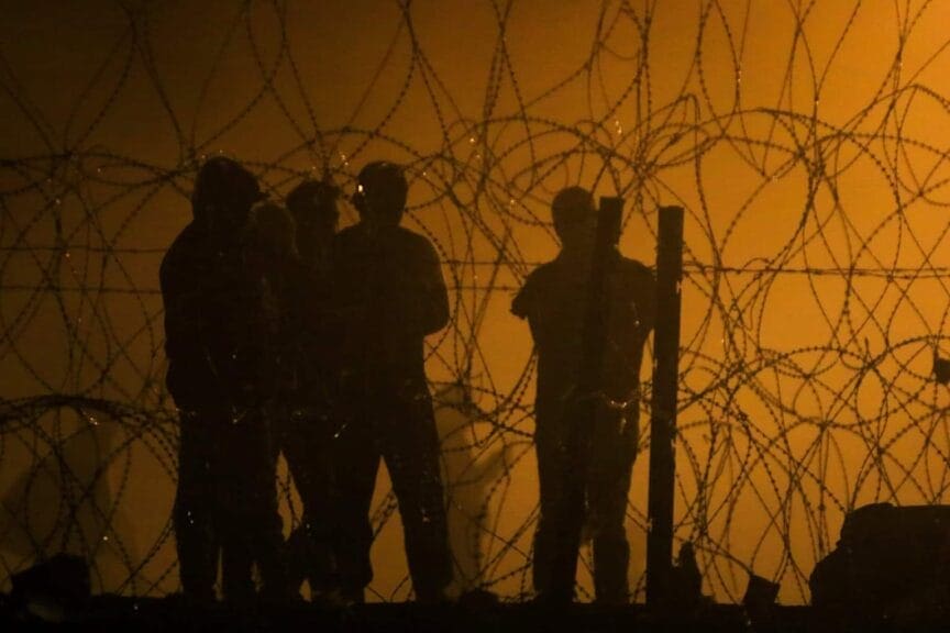 Immigrants rights migrants looking through a barbed wire fence as the sun rises in an orange sky