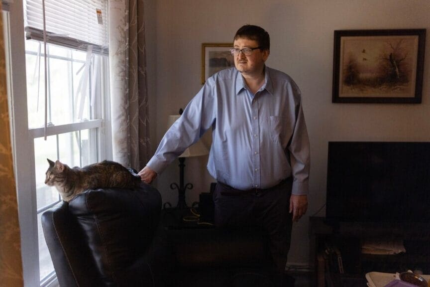 Medicaid work requirements have caused issues for this man, standing next to his cat laying on the back of an a recliner.