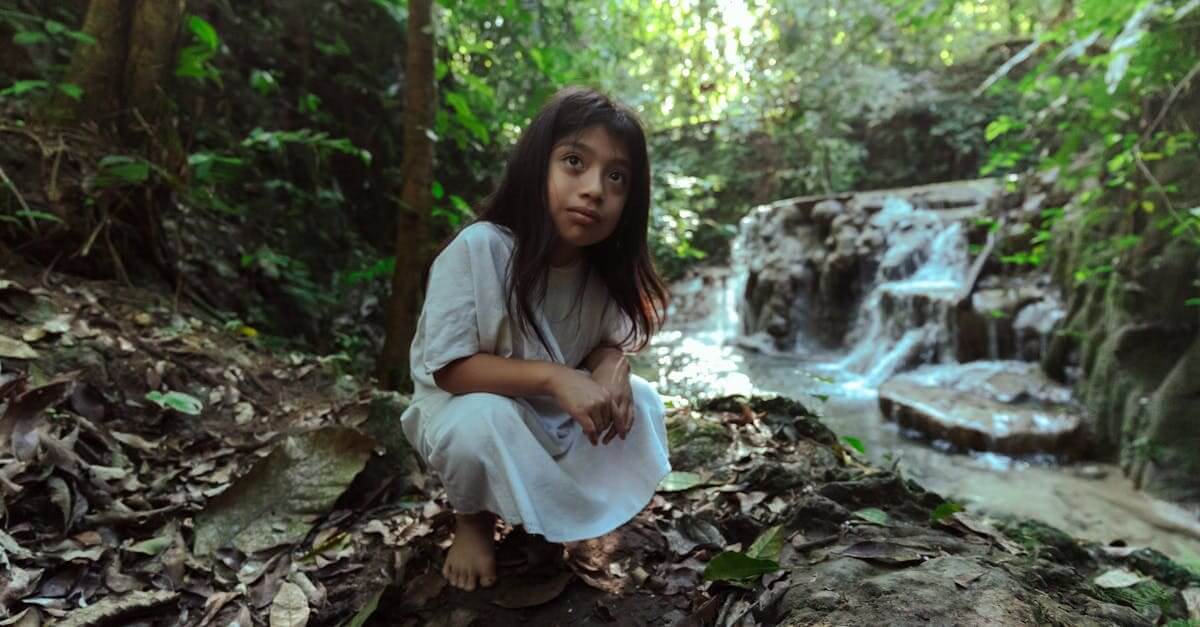 A young girl in traditional clothing explores a peaceful forest by a flowing waterfall.