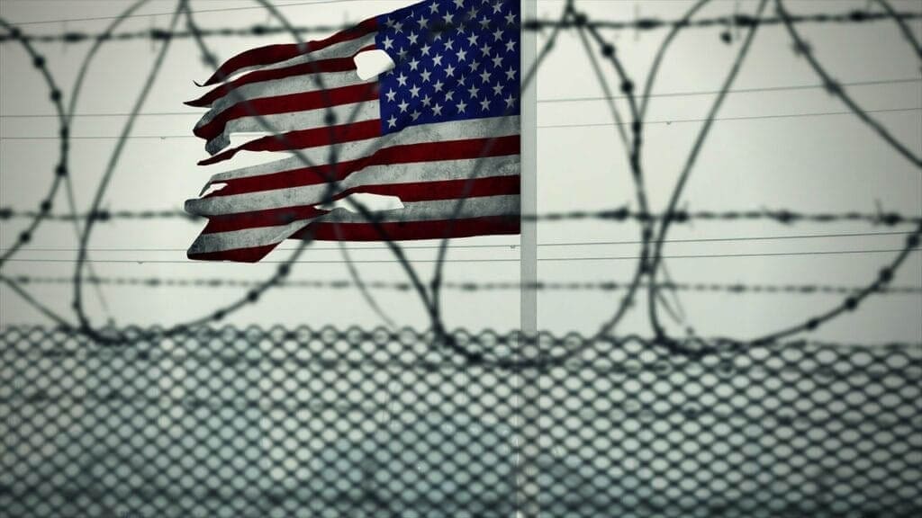 torn American flag viewed through a fence with razor wire