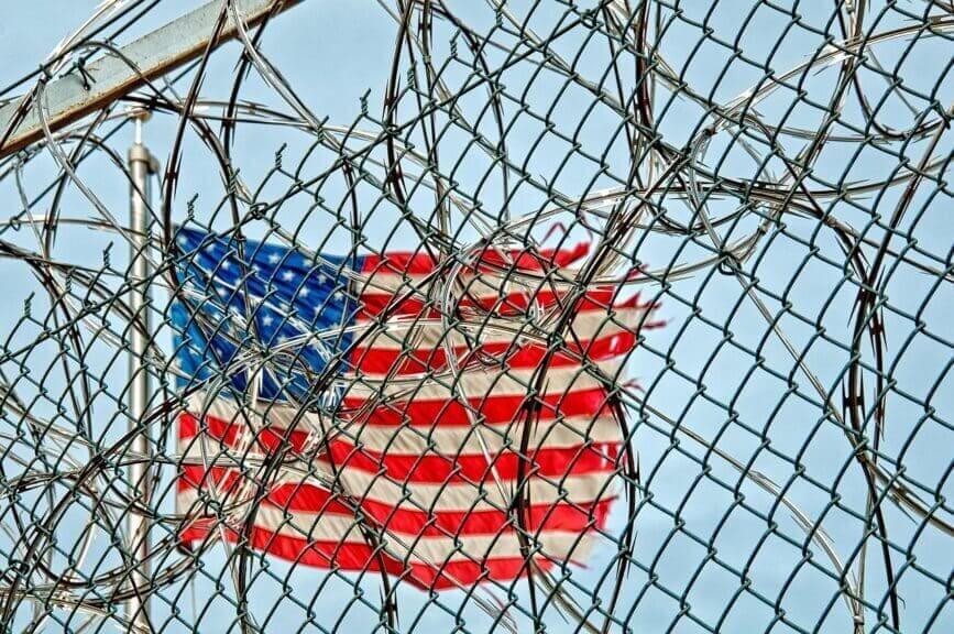 Razorwire and fencing in front of a torn American flag. 6 deaths in California ICE detention