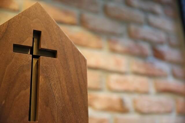 A cross carved into wood with a brick wall behind it.