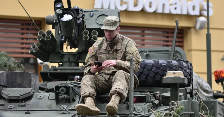A soldier in camouflage resting on an armored vehicle, using a smartphone outside a mcdonald's.